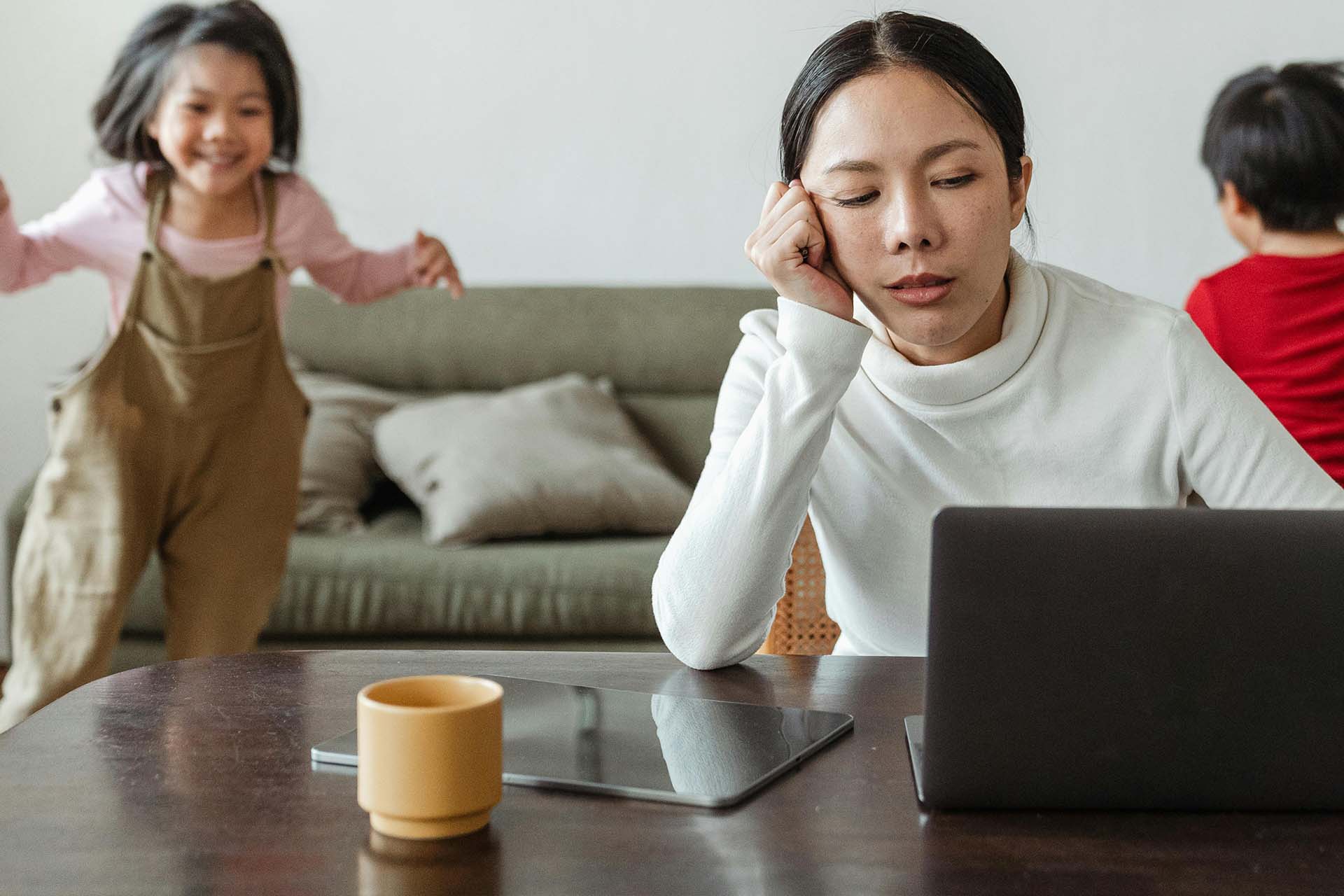 en mamma sitter trött med sin laptop och barnen leker i bakgrunden