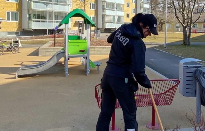 Kvinna i Fabokläder räfsar i en rabatt bredvid en lekplats.
